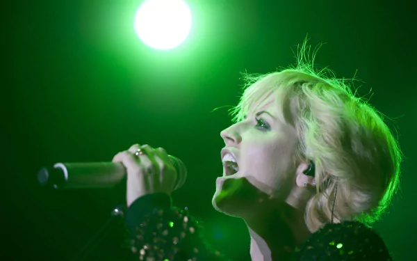 Blonde female singer mid-performance, gripping a microphone and singing under a bright green stage light — energetic live-music portrait as a 4K Ultra HD PC desktop wallpaper background
