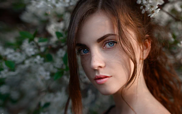 Close-up HD portrait of a redhead woman with blue eyes and freckles, surrounded by soft-focus white blossoms, creating a serene and natural desktop wallpaper background.