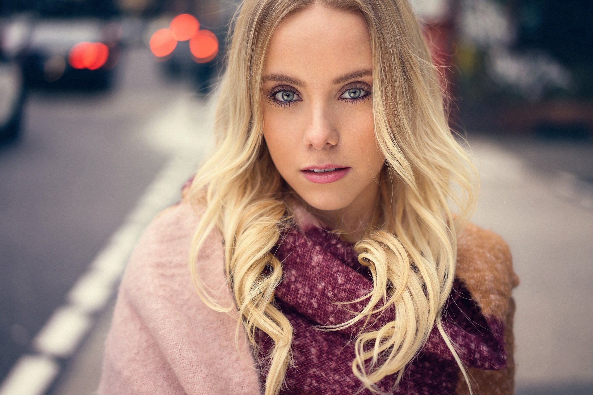 HD desktop wallpaper features a blonde model with striking blue eyes against a softly blurred bokeh city background.