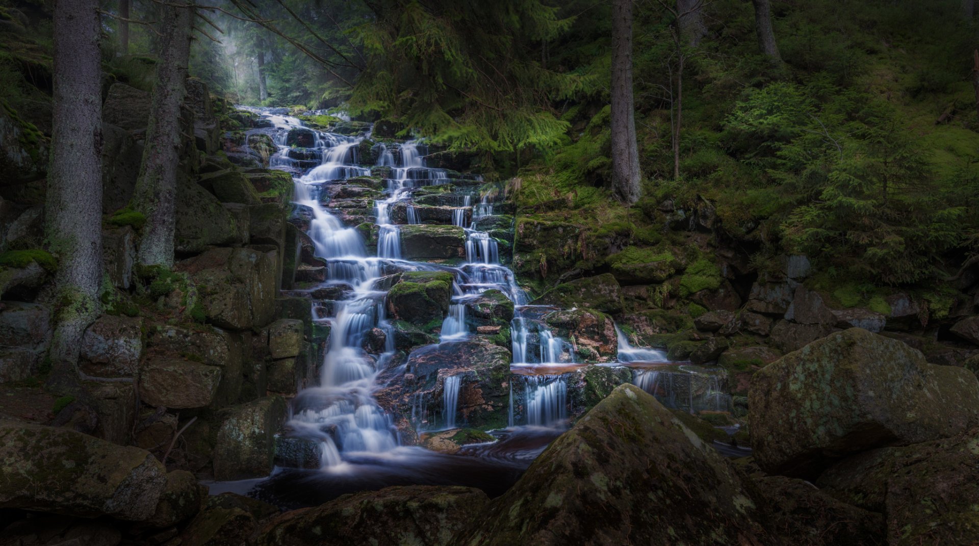 HD PC desktop wallpaper featuring a serene forest waterfall cascading over moss-covered rocks surrounded by lush greenery and tall trees.