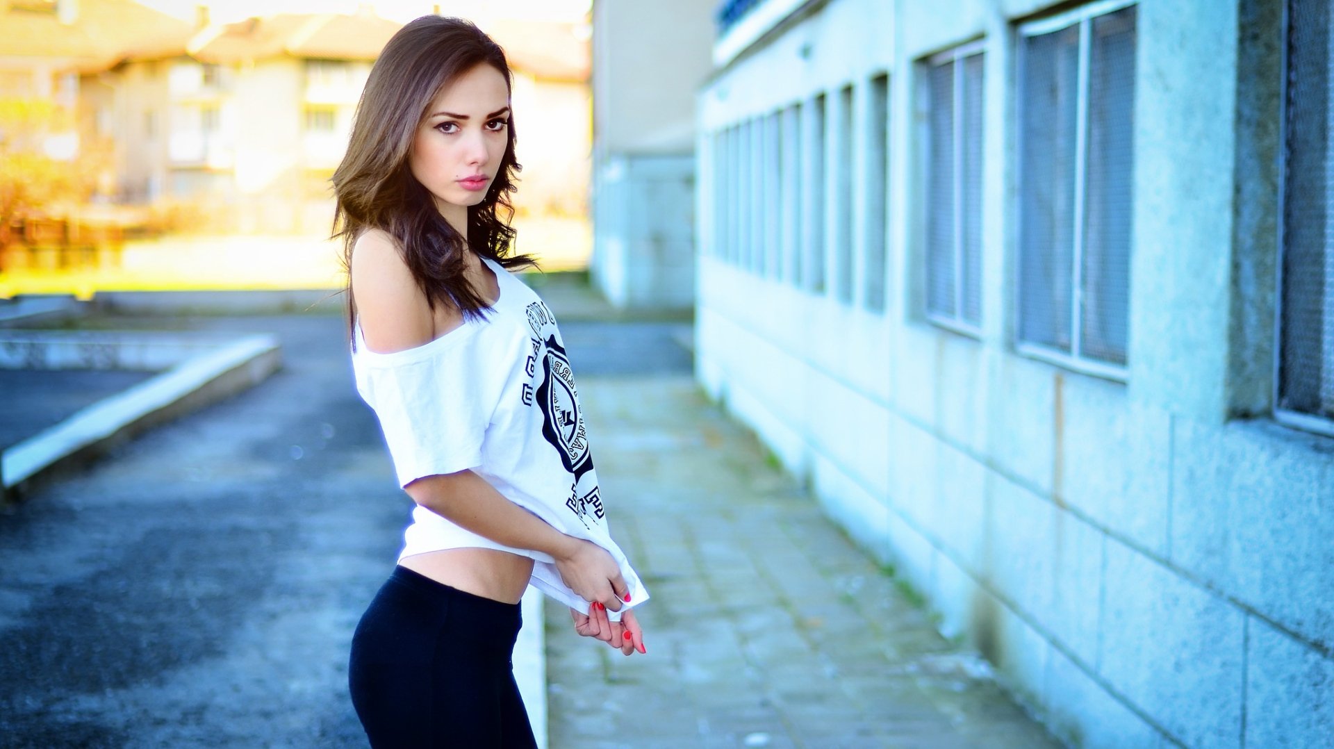 HD desktop wallpaper featuring a woman model posing outdoors next to a building, wearing a white off-shoulder top and black leggings.