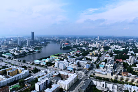 A 4K Ultra HD aerial view of Yekaterinburg, Russia, showcasing a river, city skyscrapers, roads, and man-made buildings under a partly cloudy sky.