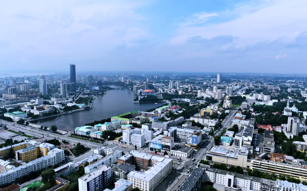 A 4K Ultra HD aerial view of Yekaterinburg, Russia, showcasing a river, city skyscrapers, roads, and man-made buildings under a partly cloudy sky.