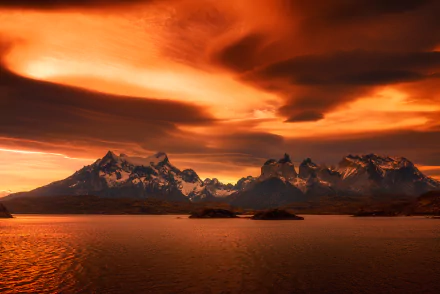 A breathtaking Patagonian sunset over Torres del Paine in Chile, featuring dramatic orange clouds reflecting on tranquil waters, creating a stunning natural landscape.