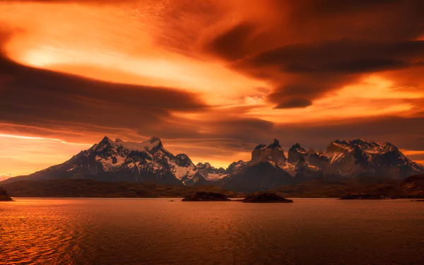 A breathtaking Patagonian sunset over Torres del Paine in Chile, featuring dramatic orange clouds reflecting on tranquil waters, creating a stunning natural landscape.