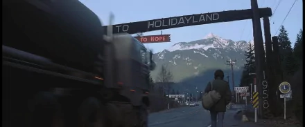 2K Quad HD PC desktop wallpaper and background of First Blood: lone backpacker walks into a mountain town under a 'TO HOLIDAYLAND / to HOPE' sign as a truck blurs past.