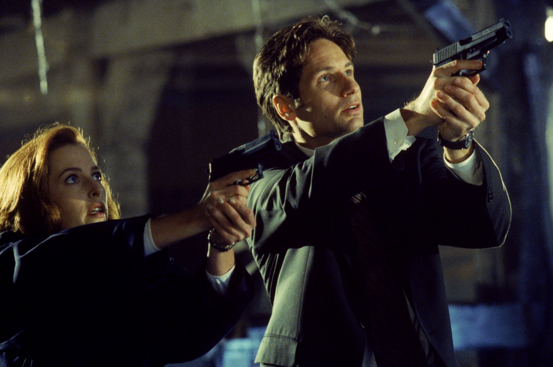 The X-Files TV show 2K Quad HD PC desktop wallpaper: two suited agents aim handguns in a dim, industrial setting, creating a tense, dramatic investigative mood.