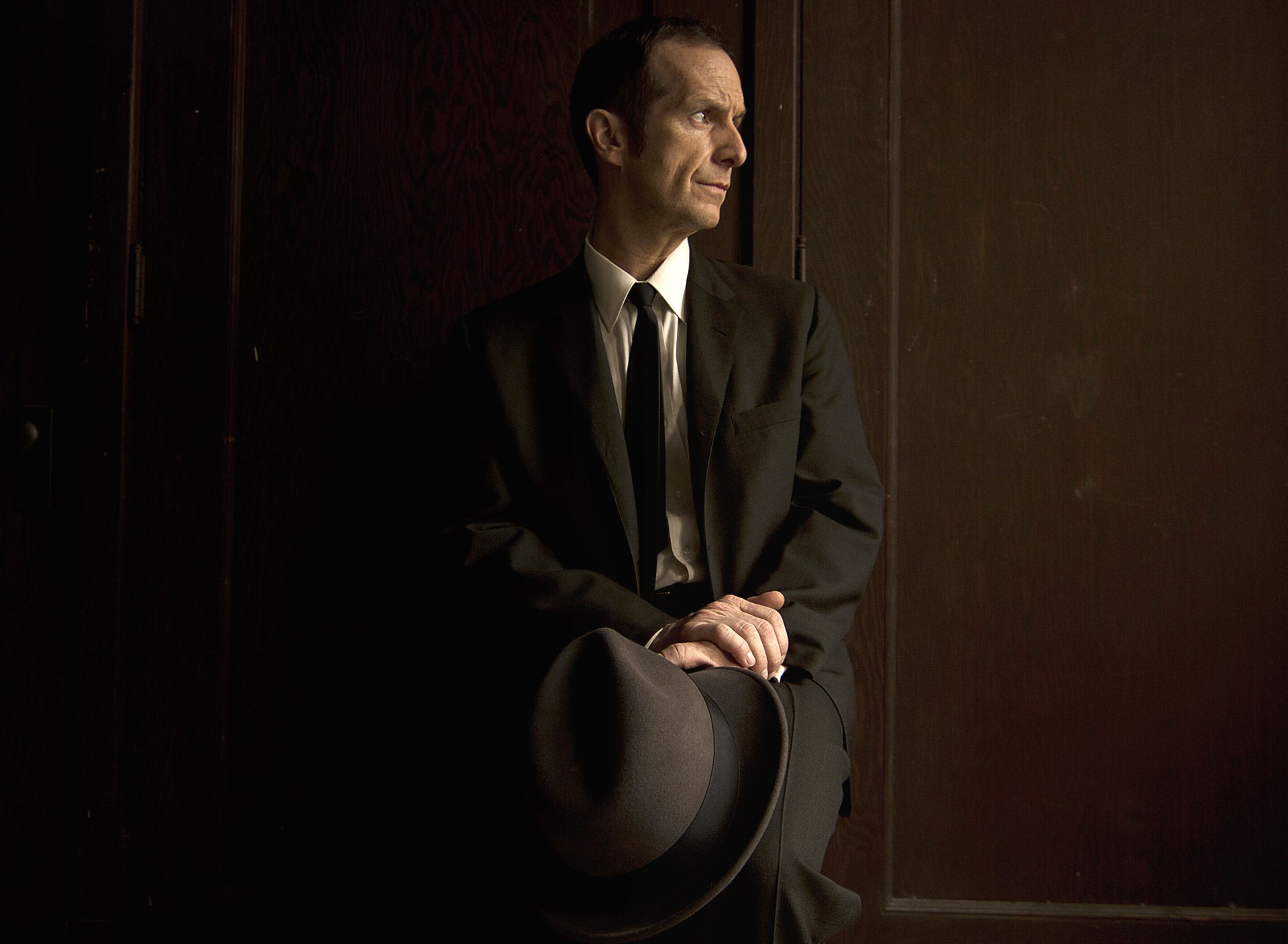 HD desktop wallpaper featuring Denis O'Hare from the TV show American Horror Story, dressed in a dark suit and holding a hat, against a moody brown background.
