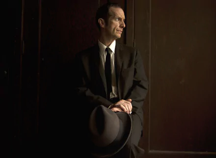 HD desktop wallpaper featuring Denis O'Hare from the TV show American Horror Story, dressed in a dark suit and holding a hat, against a moody brown background.