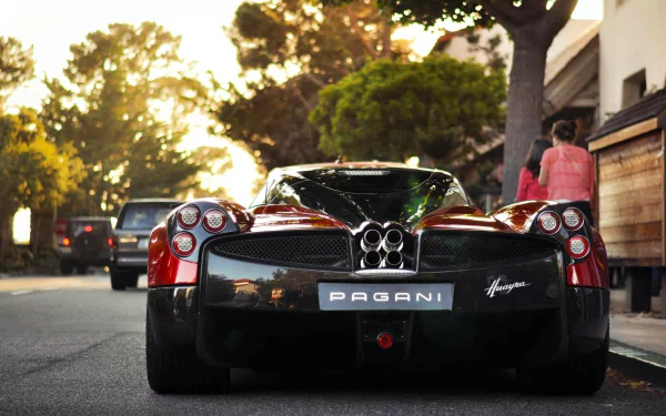 HD desktop wallpaper featuring the rear view of a sleek Pagani Huayra sports car parked on a sunlit street with trees and buildings in the background.