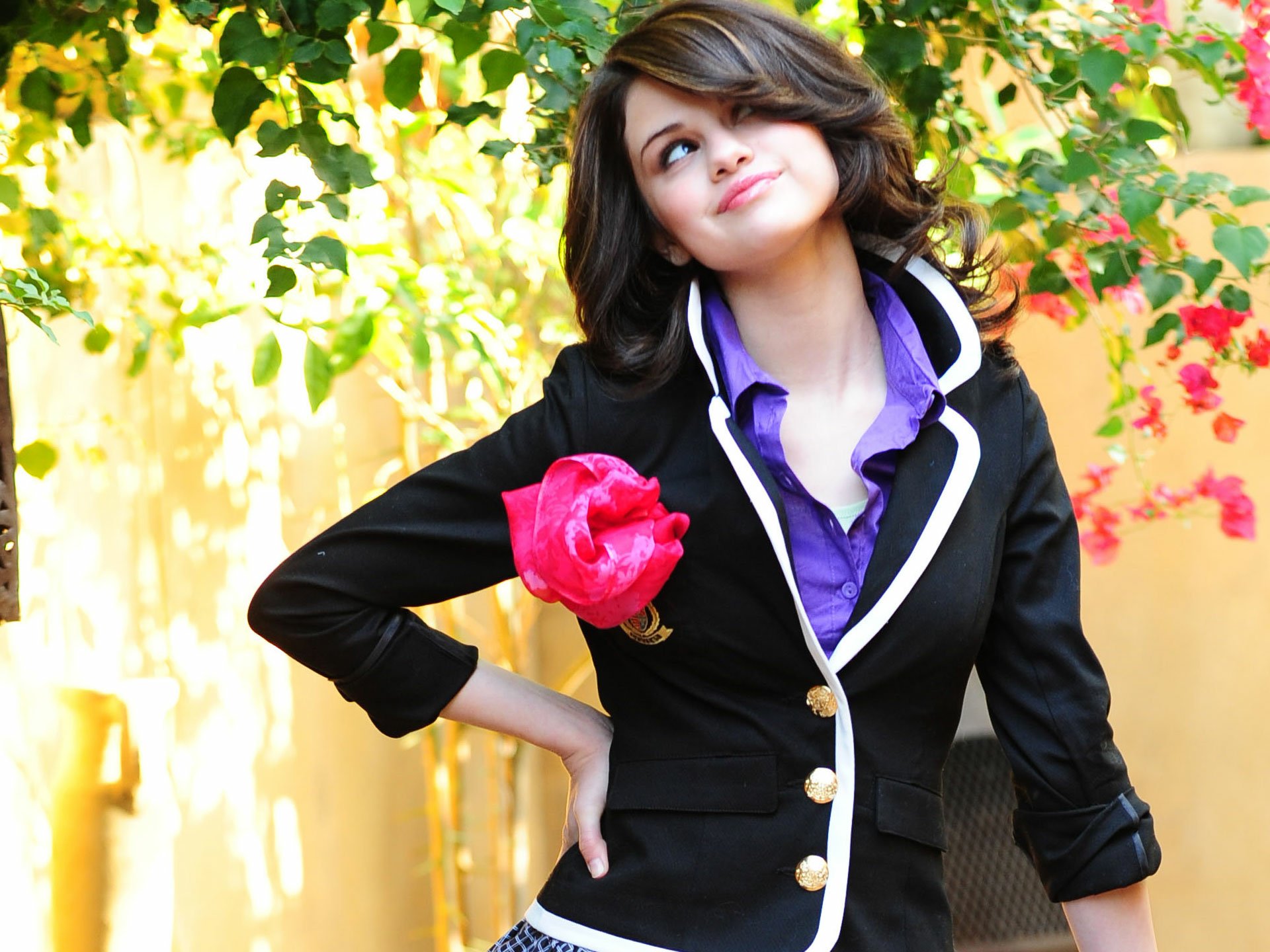 Selena Gomez poses playfully with a pink flower in a stylish black blazer, set against vibrant greenery, creating a lively music-themed HD desktop wallpaper.