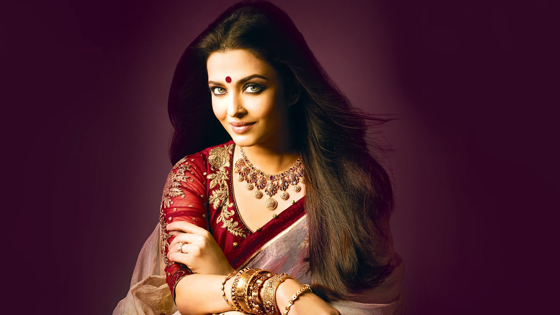 Aishwarya Rai poses elegantly in a traditional red and gold outfit, adorned with jewelry, set against a rich purple background. This captivating image serves as a stunning HD desktop wallpaper.
