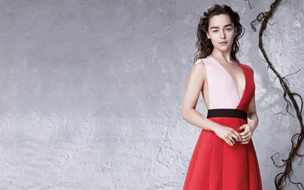 Brunette English actress Emilia Clarke poses elegantly in a striking red and pink dress against a textured backdrop, showcasing her captivating style.