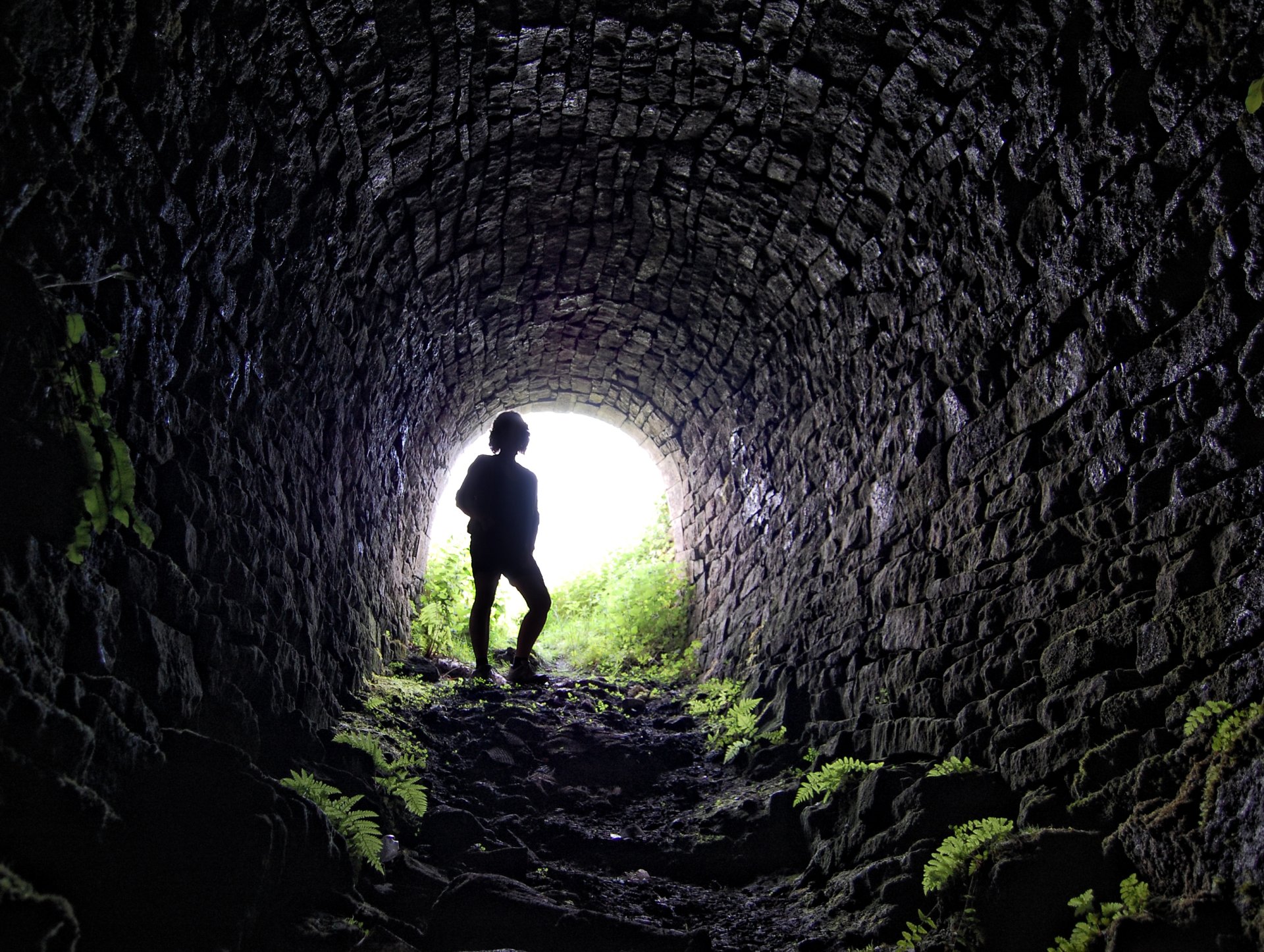 HD PC desktop wallpaper showing a silhouette of a person standing inside a man-made stone tunnel with bright light and greenery visible outside.