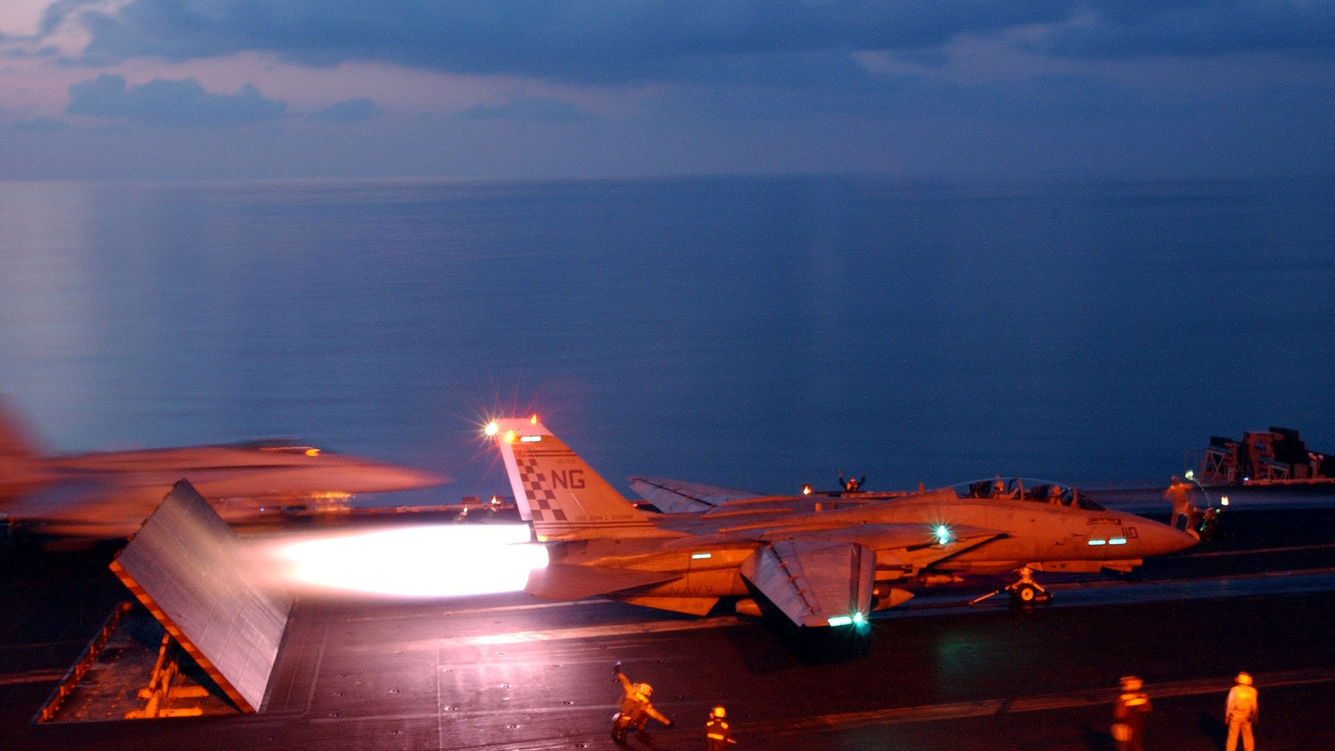 HD desktop wallpaper showing a military Grumman F-14 Tomcat jet launching from an aircraft carrier at dusk with crew members on deck.
