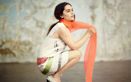 Emilia Clarke poses gracefully in a stylish outfit with an orange scarf, set against a textured backdrop. This vibrant image serves as an elegant HD wallpaper.