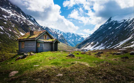 A man-made wooden cabin nestled in the mountainous landscape of Norway, surrounded by green grass and snow-capped peaks under a partly cloudy sky.
