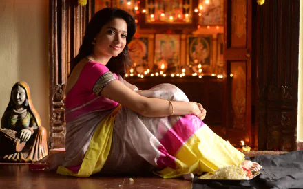 Portrait of a woman in a pink, white and yellow saree seated by a lit shrine with diyas and warm lights; 2K Quad HD PC desktop wallpaper/background.