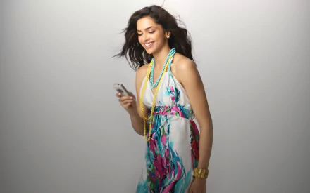 A vibrant HD wallpaper featuring Deepika Padukone gracefully smiling and texting, wearing a colorful dress and stylish accessories against a soft background.