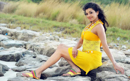 Kajal Aggarwal poses gracefully on rocky terrain, wearing a vibrant yellow outfit, set against a backdrop of lush greenery. This HD image serves as an appealing desktop wallpaper.