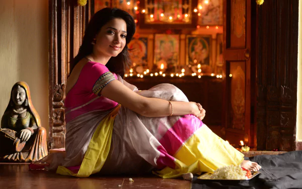 Portrait of a woman in a pink, white and yellow saree seated by a lit shrine with diyas and warm lights; 2K Quad HD PC desktop wallpaper/background.