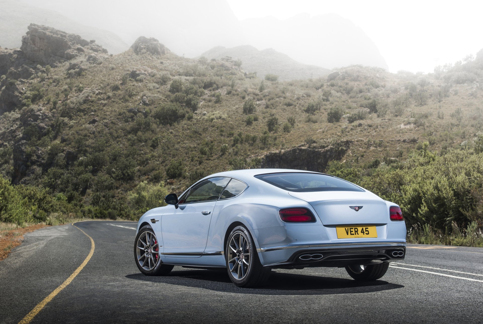 Light blue Bentley Continental GT on a winding mountain road, rear three-quarter view — 4K Ultra HD PC desktop wallpaper background.