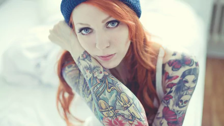HD desktop wallpaper featuring a woman with long red hair and colorful tattoos on her arms, wearing a blue beanie, looking directly at the camera.
