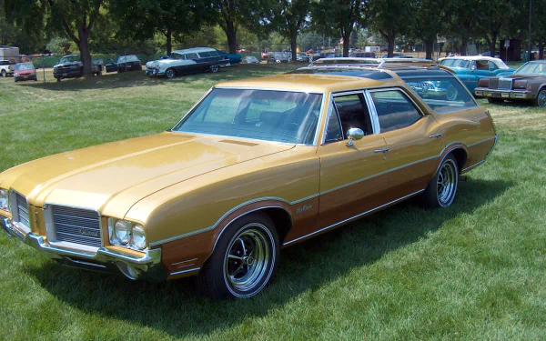 Gold Oldsmobile Vista Cruiser station wagon parked on grass at a car show, presented as an HD PC desktop wallpaper/background.