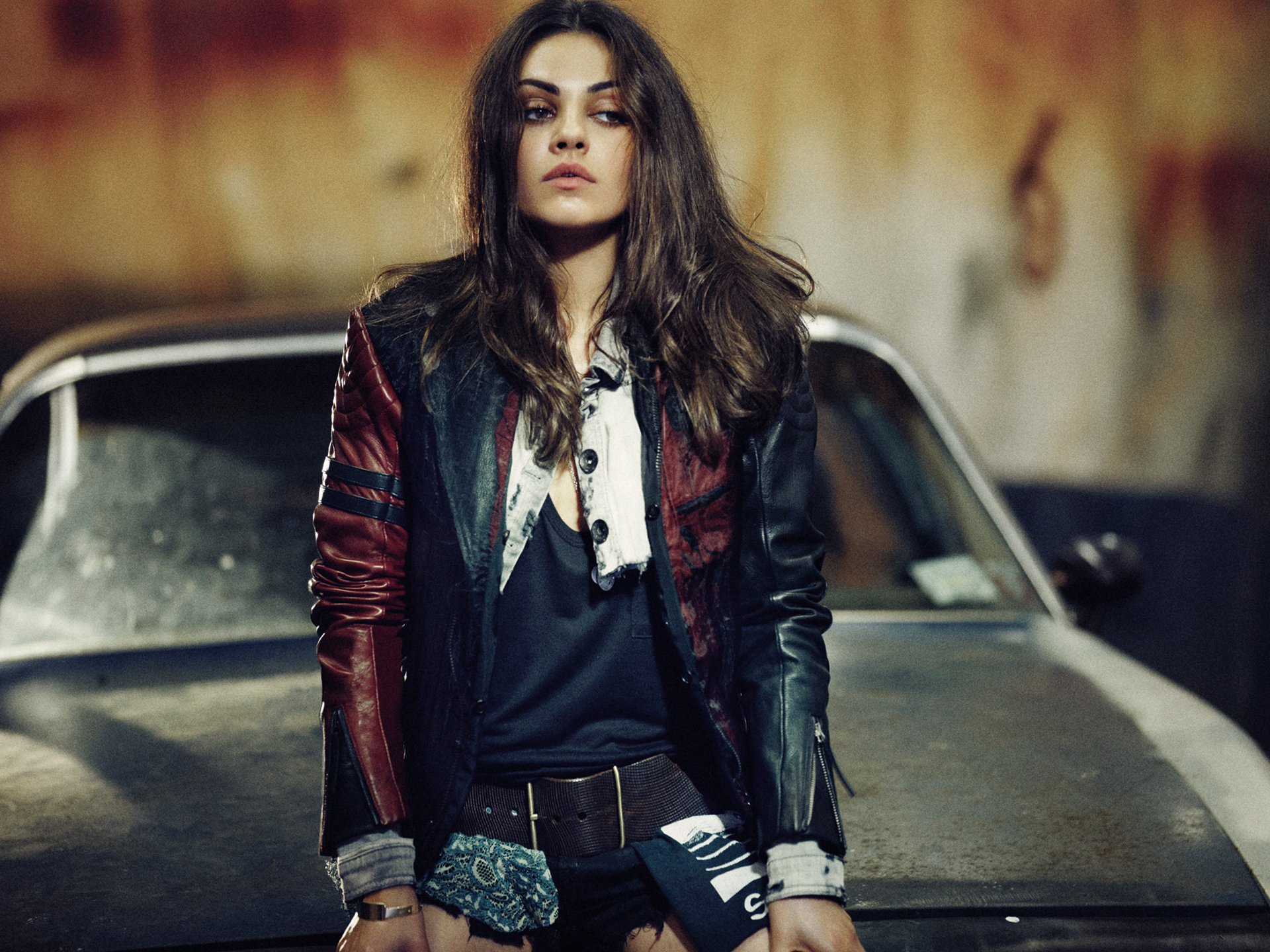 HD desktop wallpaper featuring brunette actress and celebrity Mila Kunis standing confidently in front of a weathered car with a stylish leather jacket.