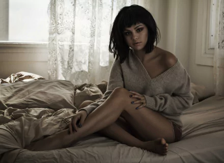 Selena Gomez is captured in a cozy scene, sitting on a bed in a soft sweater, with natural light filtering through sheer curtains, creating a serene atmosphere. HD desktop wallpaper.
