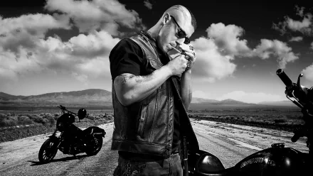 A monochrome HD wallpaper featuring a character from the TV show Sons of Anarchy, standing on a deserted road with motorcycles in the background, exuding a rugged, rebellious vibe.
