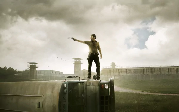 Andrew Lincoln as Rick Grimes standing on a vehicle with a revolver in a prison yard, featuring a dramatic sky and bleak landscape. HD wallpaper from the TV show The Walking Dead.