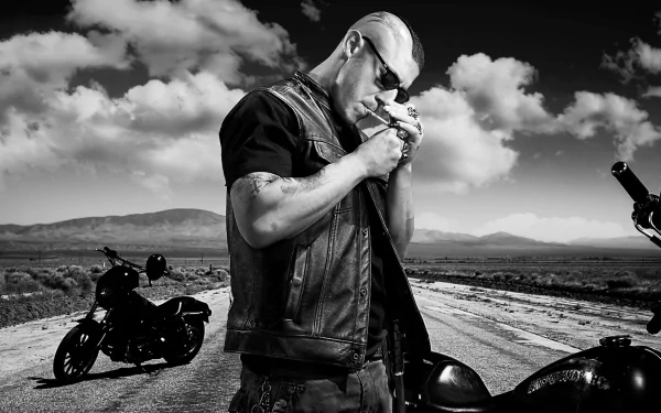 A monochrome HD wallpaper featuring a character from the TV show Sons of Anarchy, standing on a deserted road with motorcycles in the background, exuding a rugged, rebellious vibe.