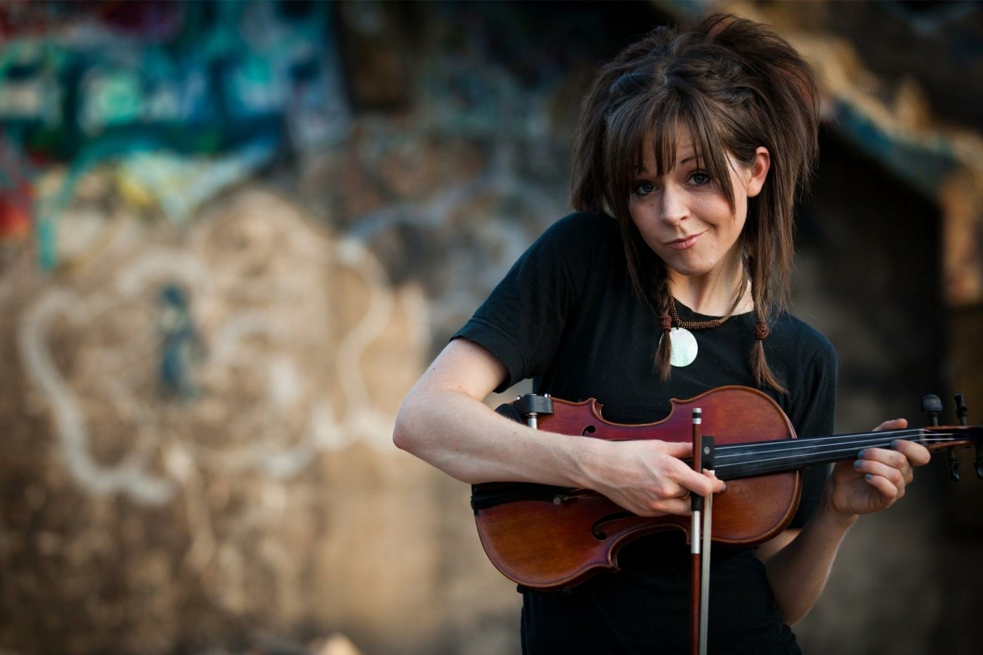 HD PC desktop wallpaper of Lindsey Stirling playing the violin, set against a blurred graffiti-covered wall, capturing a music-themed artistic vibe.
