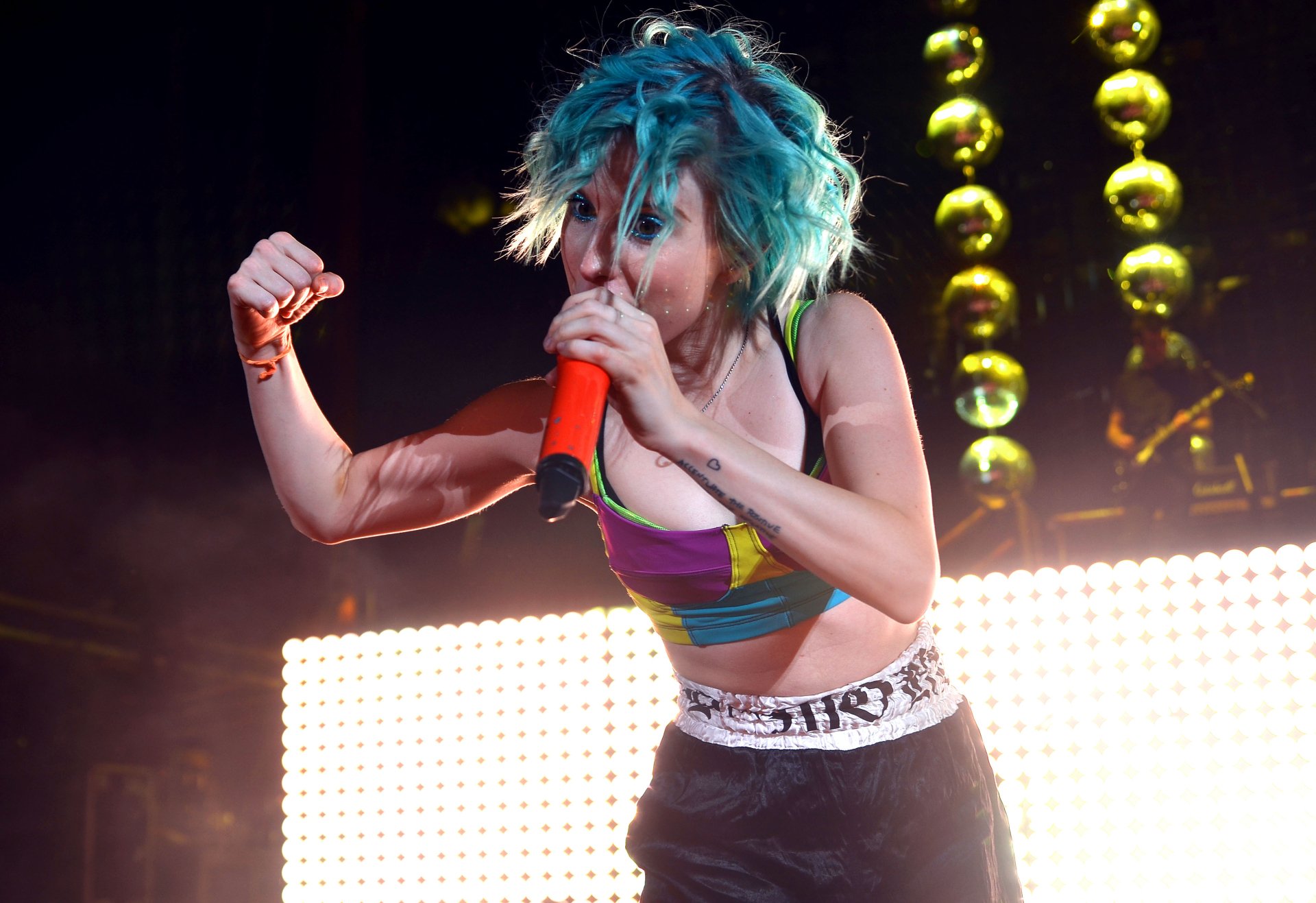 Energetic female rock singer with teal hair holding an orange mic amid stage lights — music; 2K Quad HD PC desktop wallpaper background.