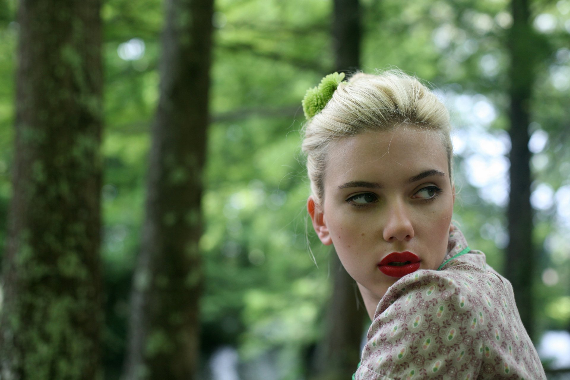 A blonde actress poses with vibrant lipstick against a softly blurred green backdrop, capturing a moment of elegance and allure in this 4K Ultra HD image.