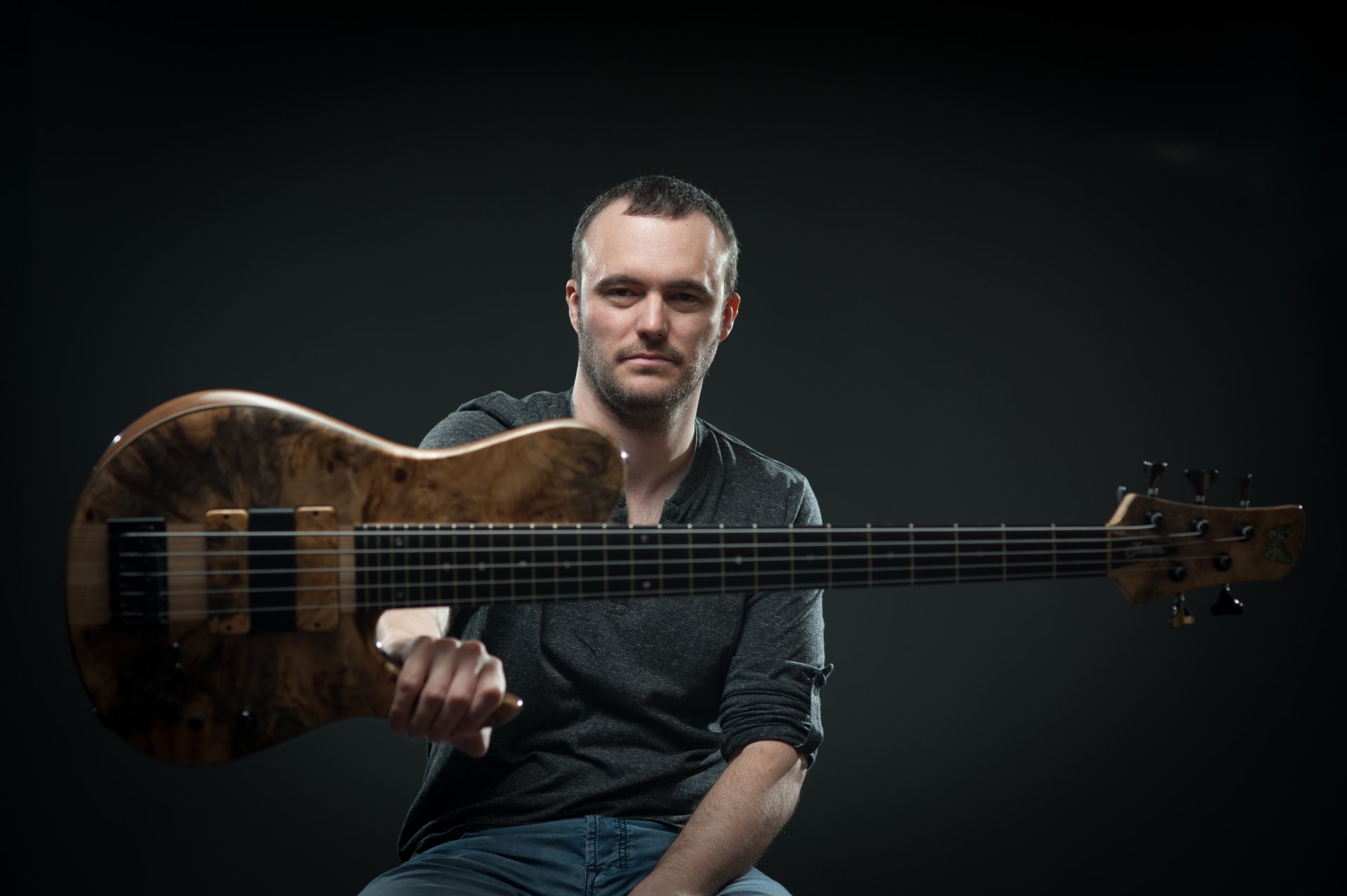 4K Ultra HD PC desktop wallpaper of a seated man against a dark background holding a long-neck electric bass guitar toward the viewer.