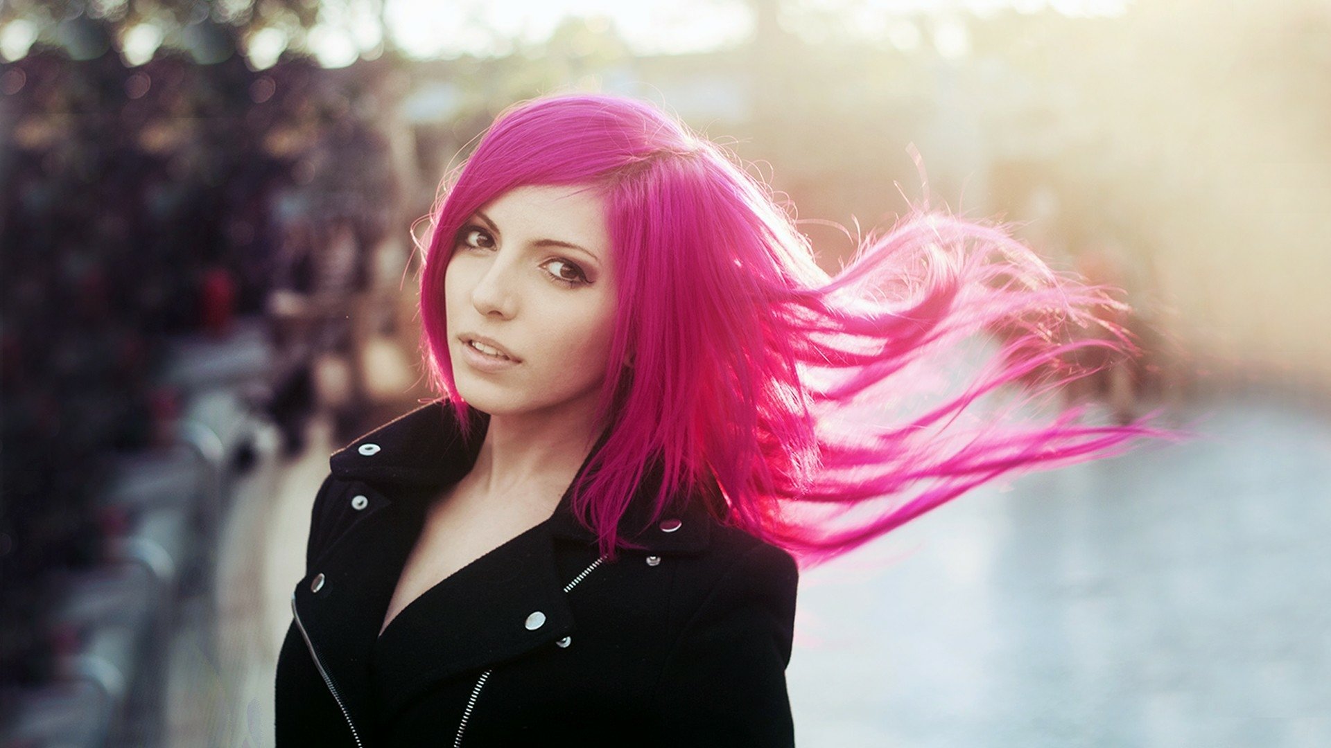 HD PC desktop wallpaper of a model woman with vibrant pink hair blowing in the wind, wearing a black coat against a softly lit urban background.