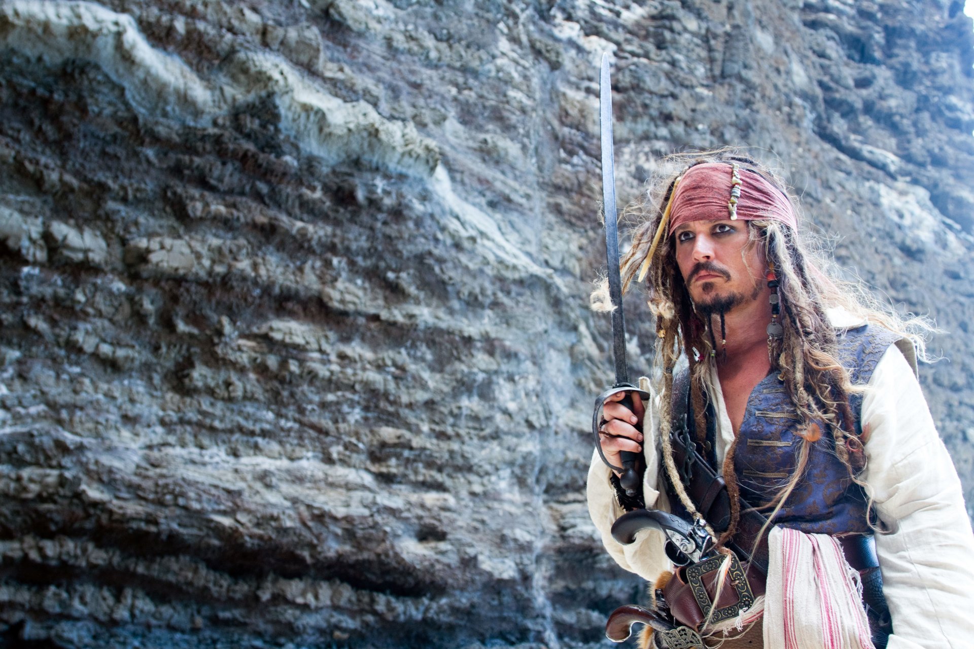 Johnny Depp as Jack Sparrow from Pirates of the Caribbean: On Stranger Tides, standing with a sword against a rocky cliff, in a 4K Ultra HD desktop wallpaper.