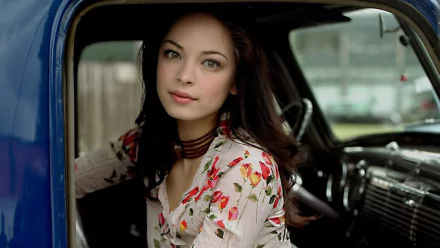 HD PC desktop wallpaper featuring celebrity Kristin Kreuk sitting inside a vintage car, wearing a floral top and looking directly at the camera.