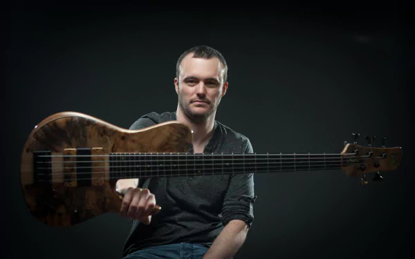 4K Ultra HD PC desktop wallpaper of a seated man against a dark background holding a long-neck electric bass guitar toward the viewer.