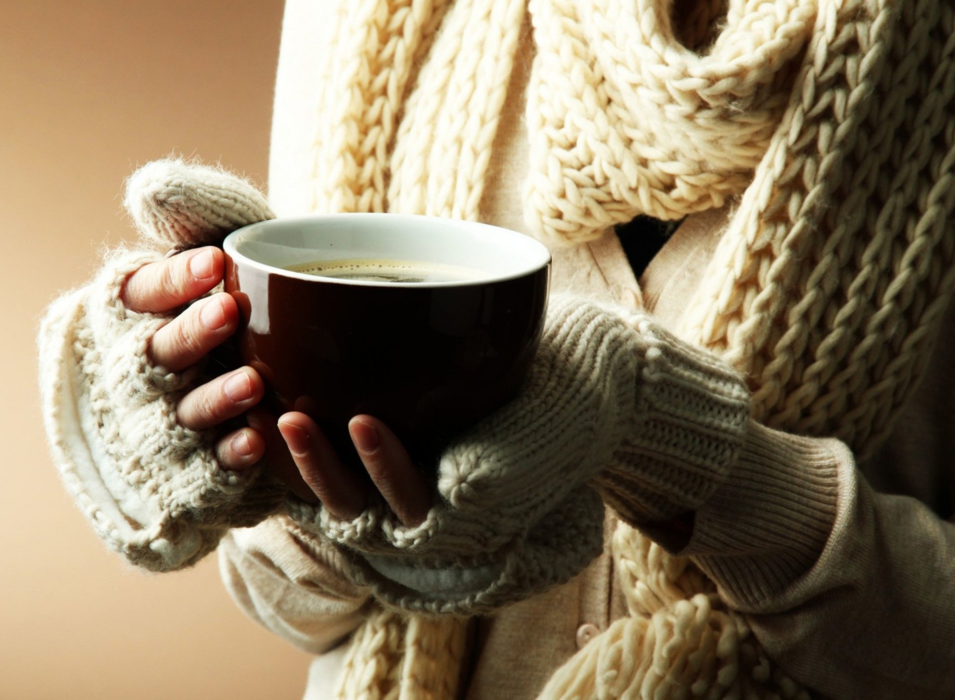 A hand wearing cozy knit gloves holds a steaming cup of coffee, set against a softly blurred background, captured in HD for a warm desktop wallpaper.