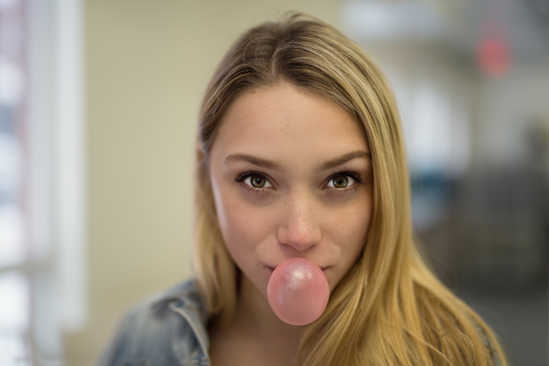 Close-up of a blonde woman with green eyes and freckles, chewing bubblegum, captured in a moody 4K Ultra HD desktop wallpaper.