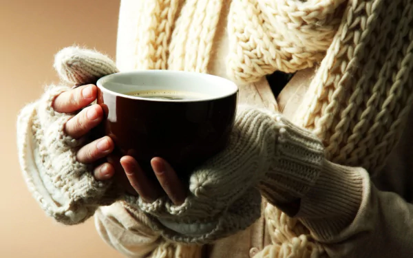A hand wearing cozy knit gloves holds a steaming cup of coffee, set against a softly blurred background, captured in HD for a warm desktop wallpaper.