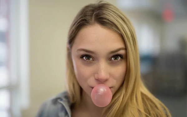 Close-up of a blonde woman with green eyes and freckles, chewing bubblegum, captured in a moody 4K Ultra HD desktop wallpaper.