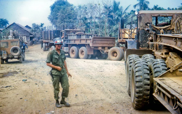 HD PC desktop wallpaper showing a Vietnam War military scene with a soldier walking near trucks and equipment in a rural setting.