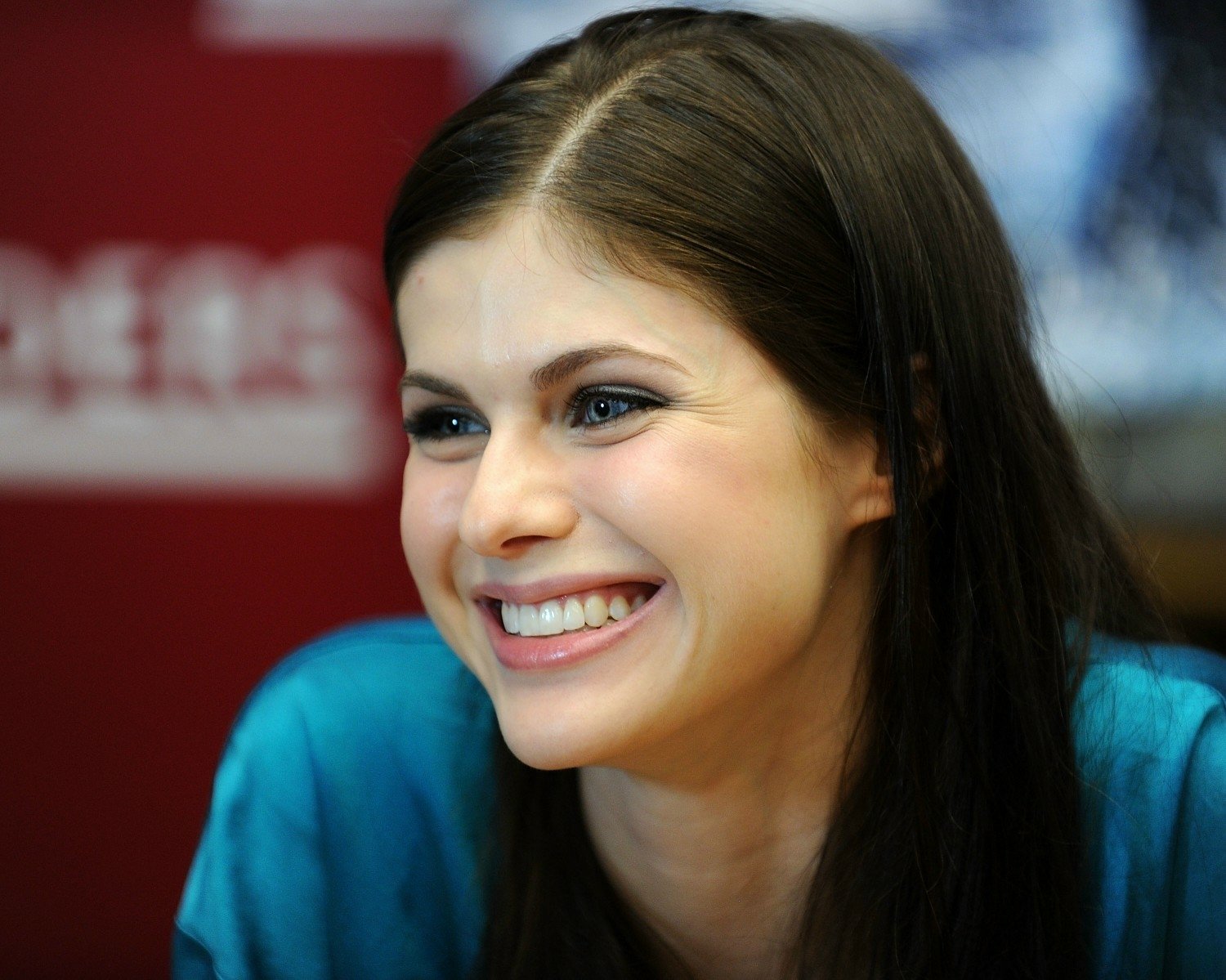 A bright and cheerful portrait of Alexandra Daddario, showcasing her joyful smile, set against a simple red background. This HD image serves as a striking desktop wallpaper.