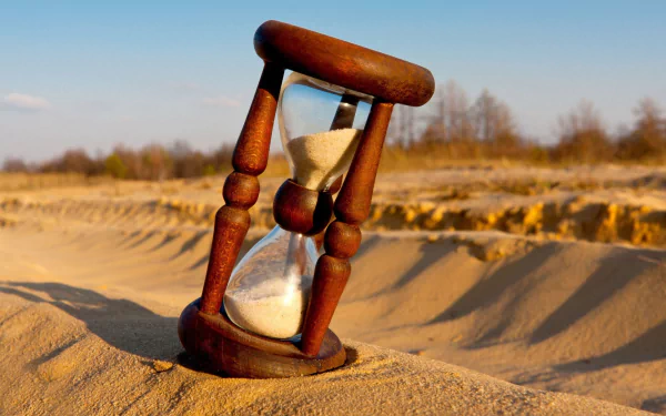 A wooden hourglass sits on sandy terrain, with fine grains cascading through it. This 4K Ultra HD wallpaper captures a tranquil moment of time.
