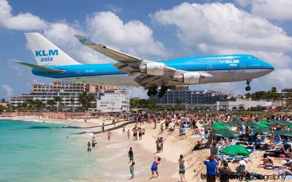 A Boeing 747 approaches Maho Beach in Saint Martin, flying low over sunbathers and umbrellas, showcasing the vibrant beach atmosphere against a clear blue sky.