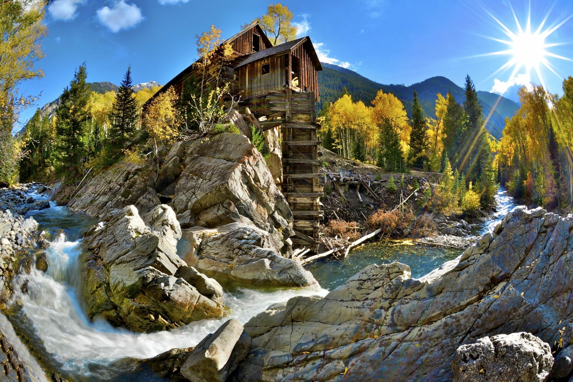 HD desktop wallpaper showcasing the man-made Crystal Mill perched on rocky terrain beside a flowing river, surrounded by autumn trees and a bright sunburst in a mountain landscape.